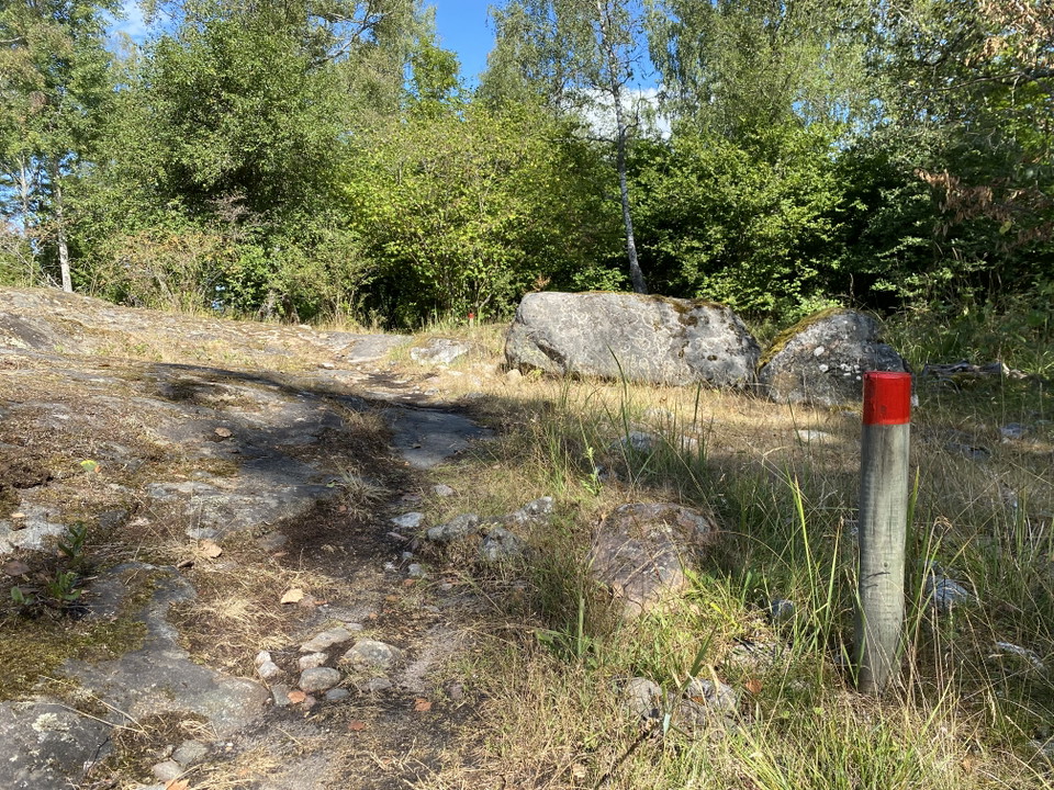 En stolpe med rödmålad topp står vid en stenig stig. Gröna träd i bakgrunden.