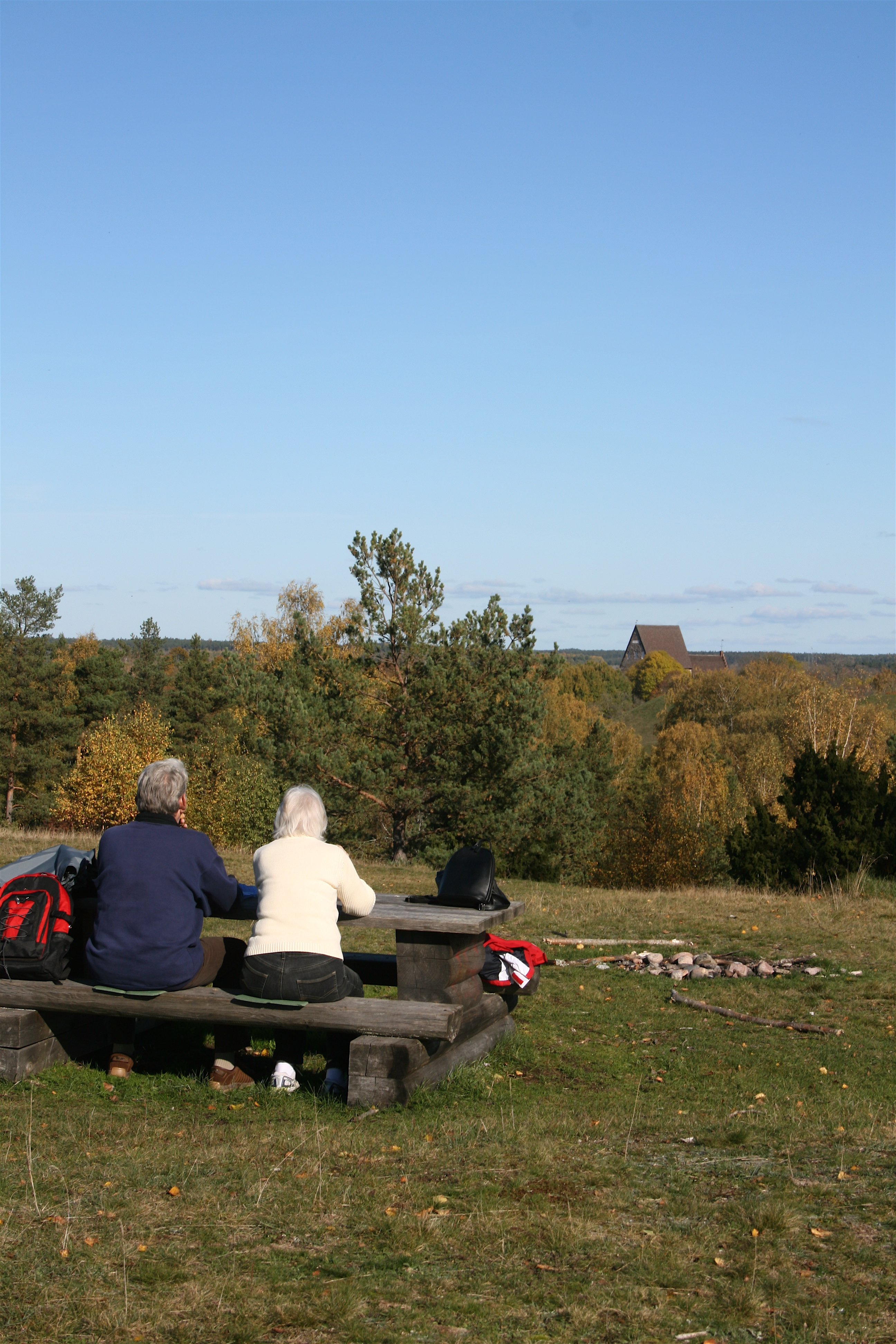 Ett äldre par sitter vid ett bänkbord uppe på en höjd. De blickar ut över träd och en högre byggnad i bakgrunden.