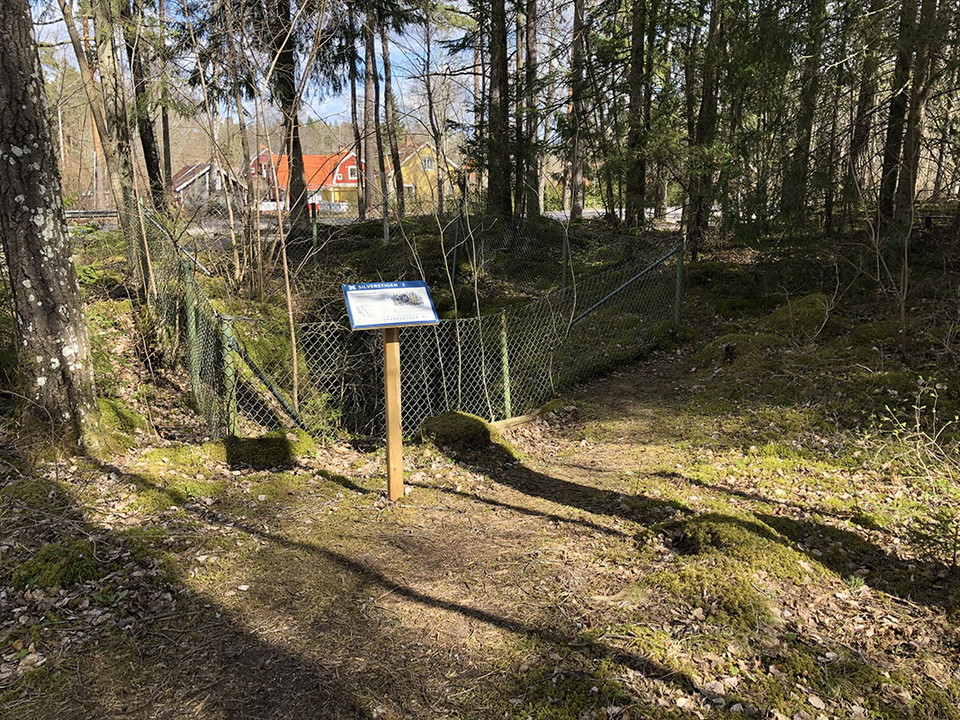 Informationstavla vid inhägnat gammalt hål i marken. I bakgrunden syns hustak.