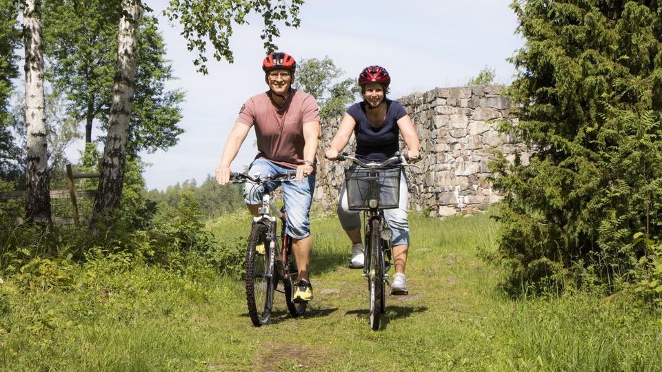 Två personer cyklar på en somrig skogsväg med en stenlagd ruin i bakgrunden.