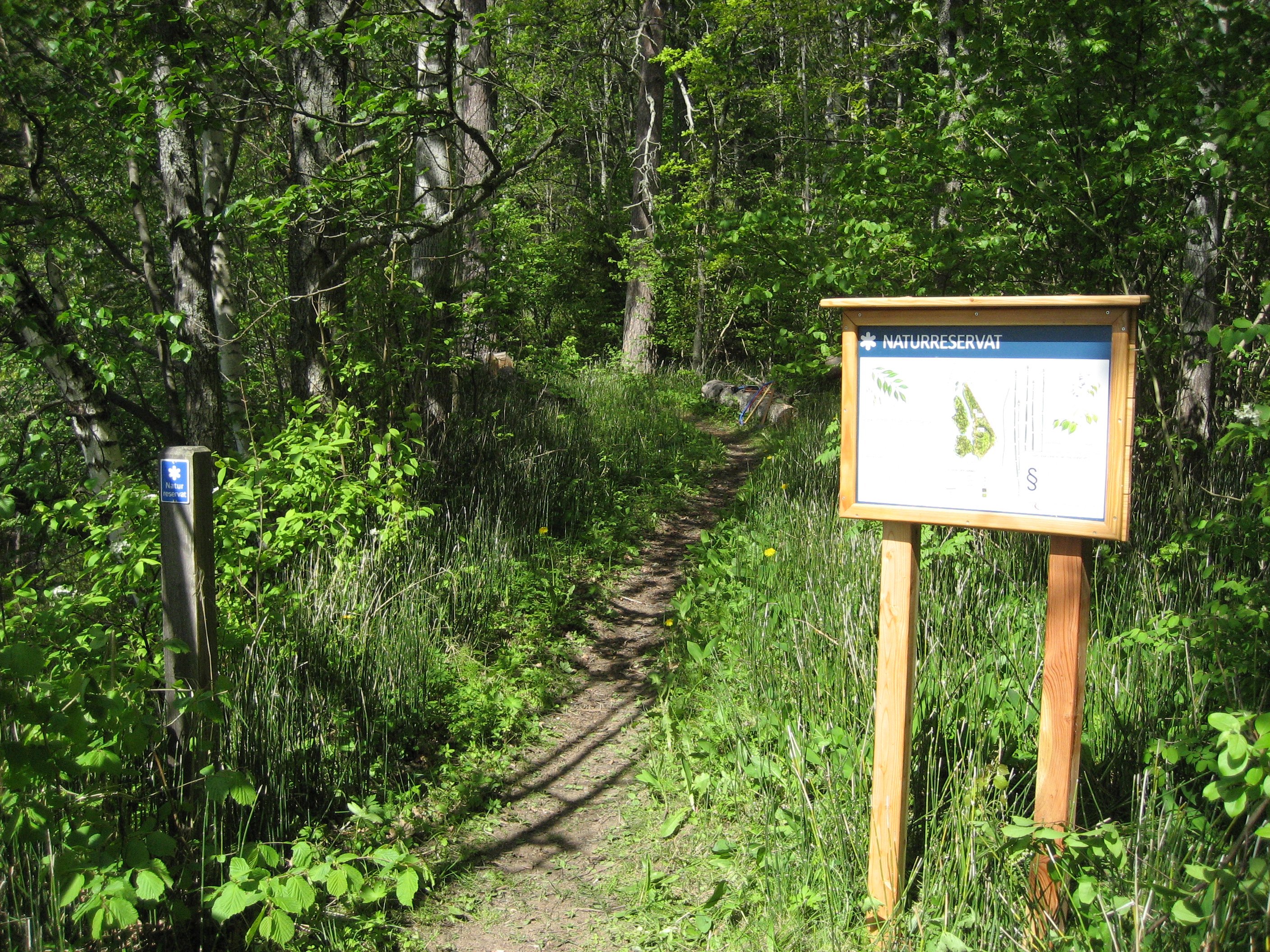 Vid en smal skogsstig står en informationsskylt och en stigmarkerning med skylten "Naturreservat". Det är tät skog på båda sidorna av stigen.