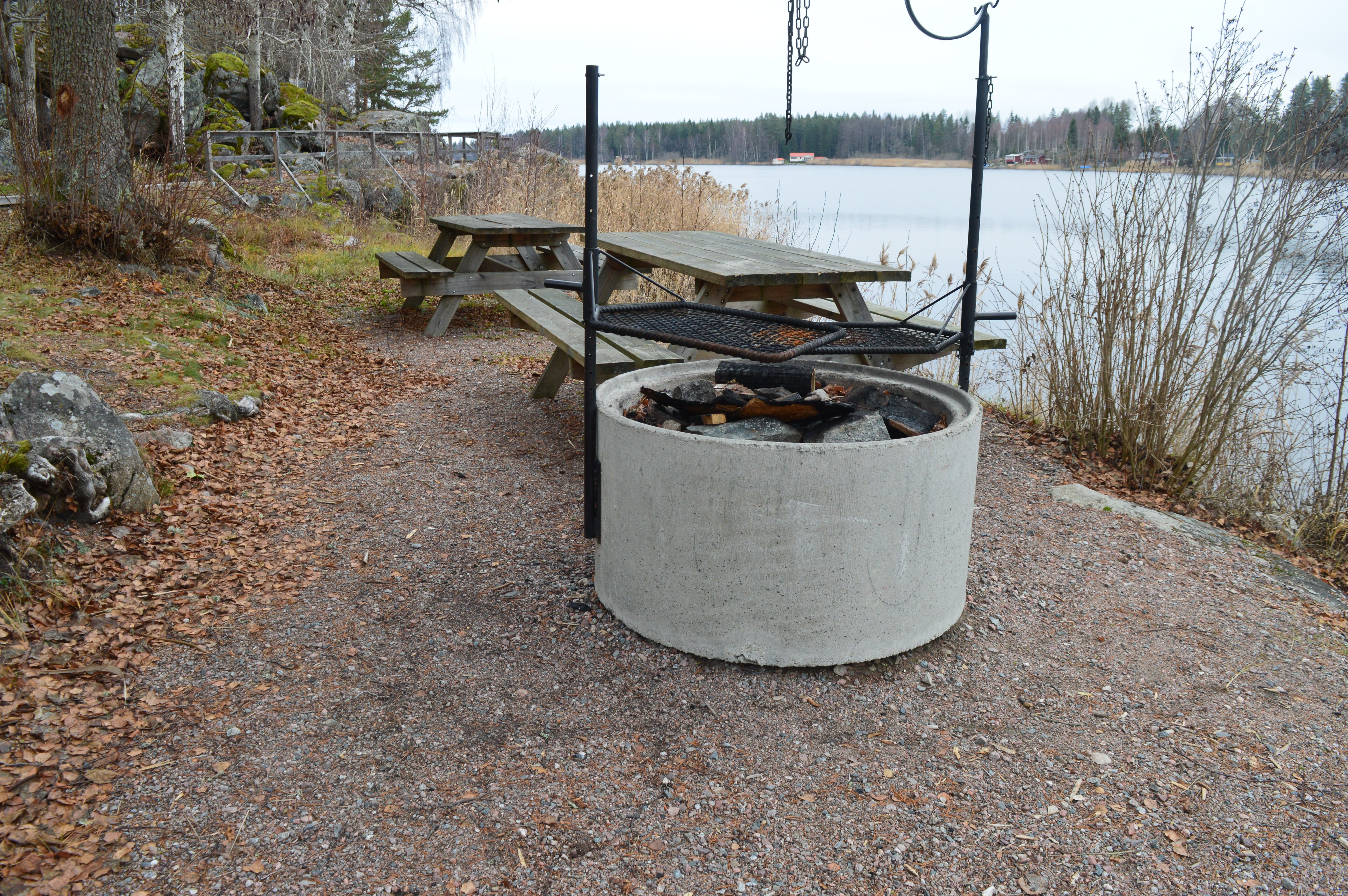 En grillplats med två grillgaller ovanpå står på grusad yta vid vattnet. Bakom grillplatser finns två bänkbord.