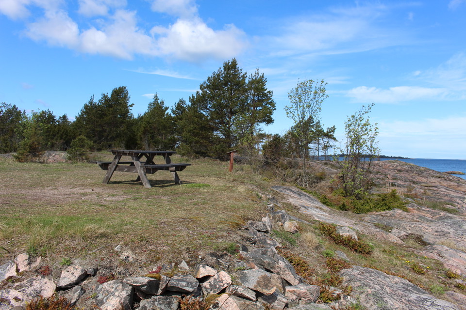På en öppen gräsyta står ett bänkbord. Nedanför rastplatsen är det klippor och hav.