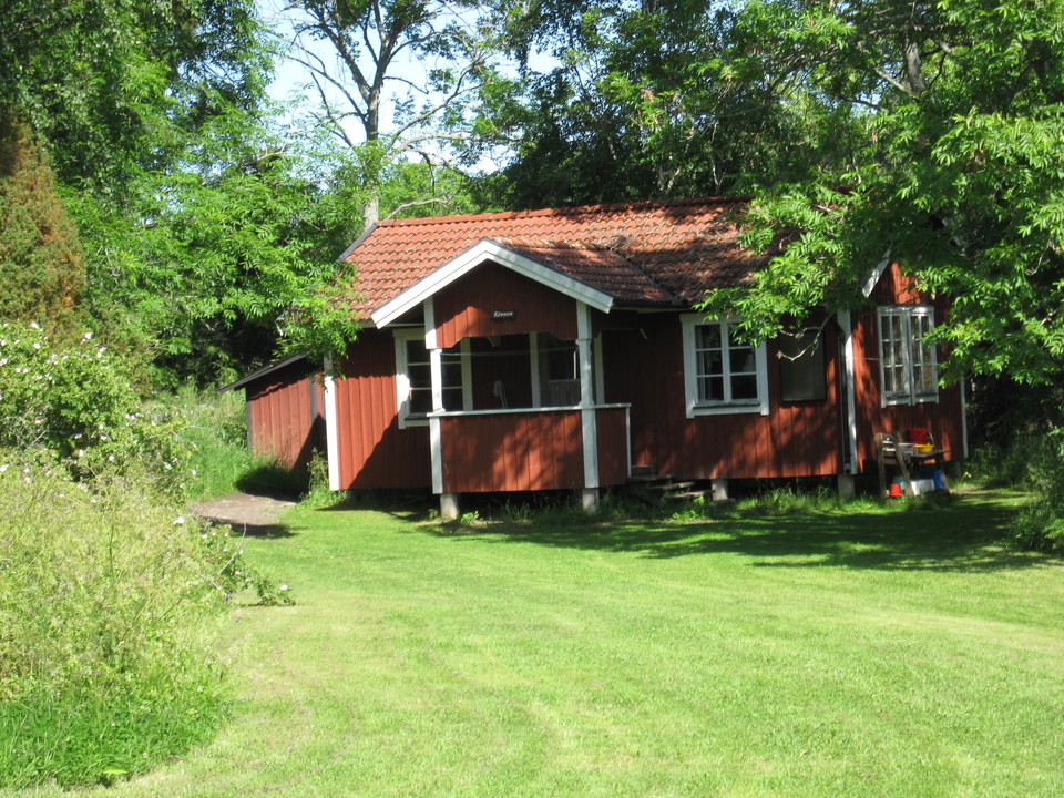 En stuga med gräsplan framför och skog bakom sig.