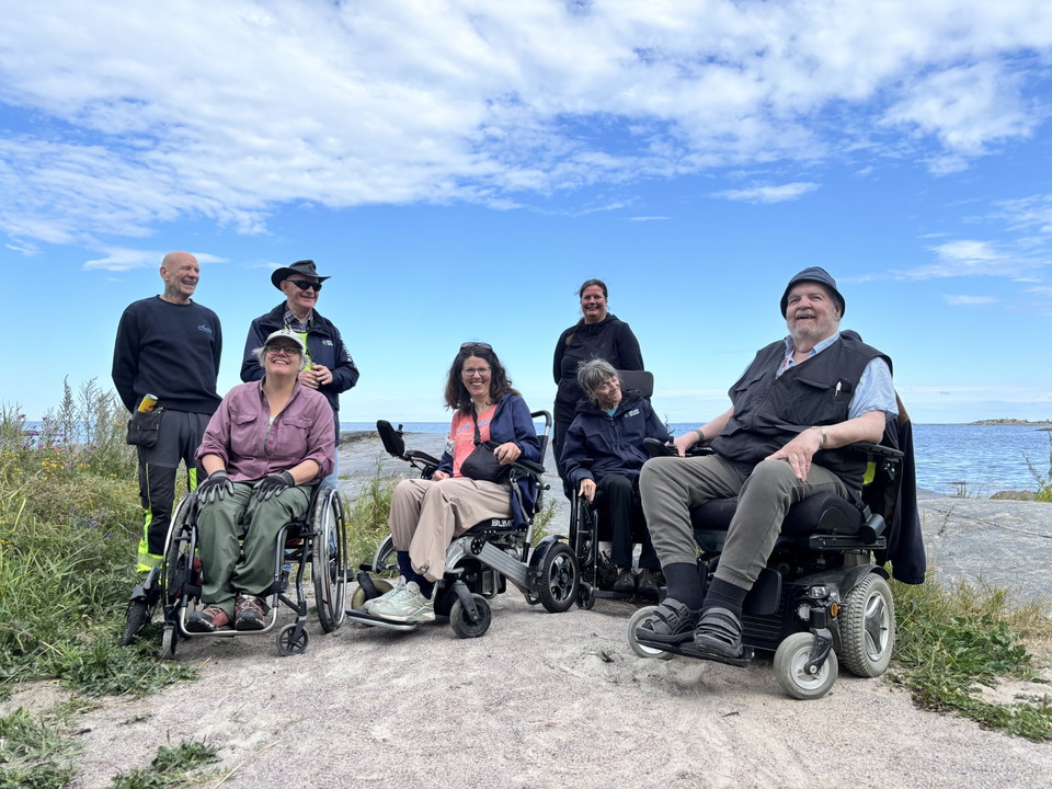 En grupp personer, några i rullstol, på en grusplan vid havet.