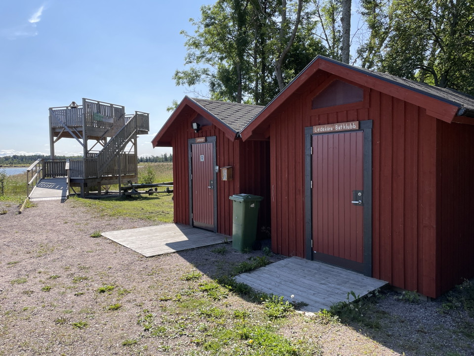 Två röda byggnader med en grusad gångväg framför. I bakgrunden syns ett fågeltorn.