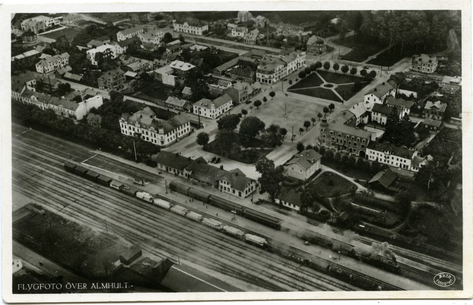 Flygfoto över Stortorget i Älmhult under tidigt 1930-tal. Ett stort torg med hus i flera våningar runt omkring och järnvägsspår intill.