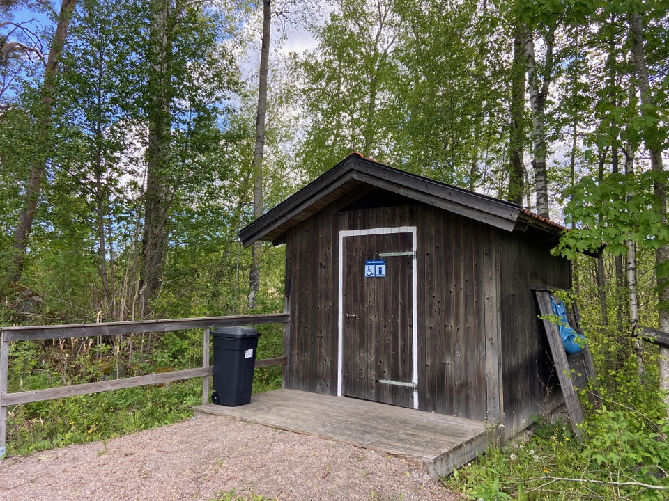 En brun byggnad med vitt foder runt dörren. En grusad gång leder fram till en kort träramp upp till byggnaden. Runt omkring står gröna träd.