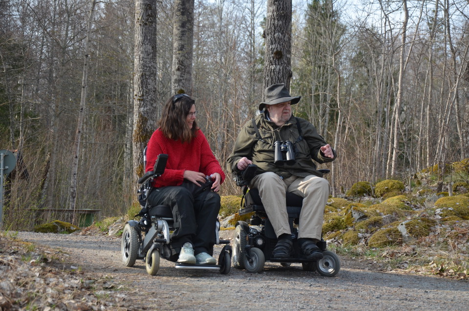 En man och kvinna i rullstol är ute på en smal grusväg i naturen. 