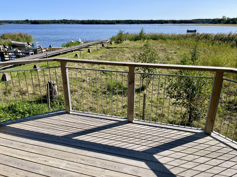 Ett trägolv med ett staket som når upp till midjehöjd. På andra sidan staketet syns en grön strandäng och i bakgrunden blått vatten.