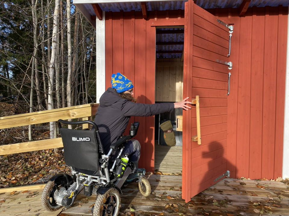 En kvinna i rullstol öppnar dörr på litet hus.