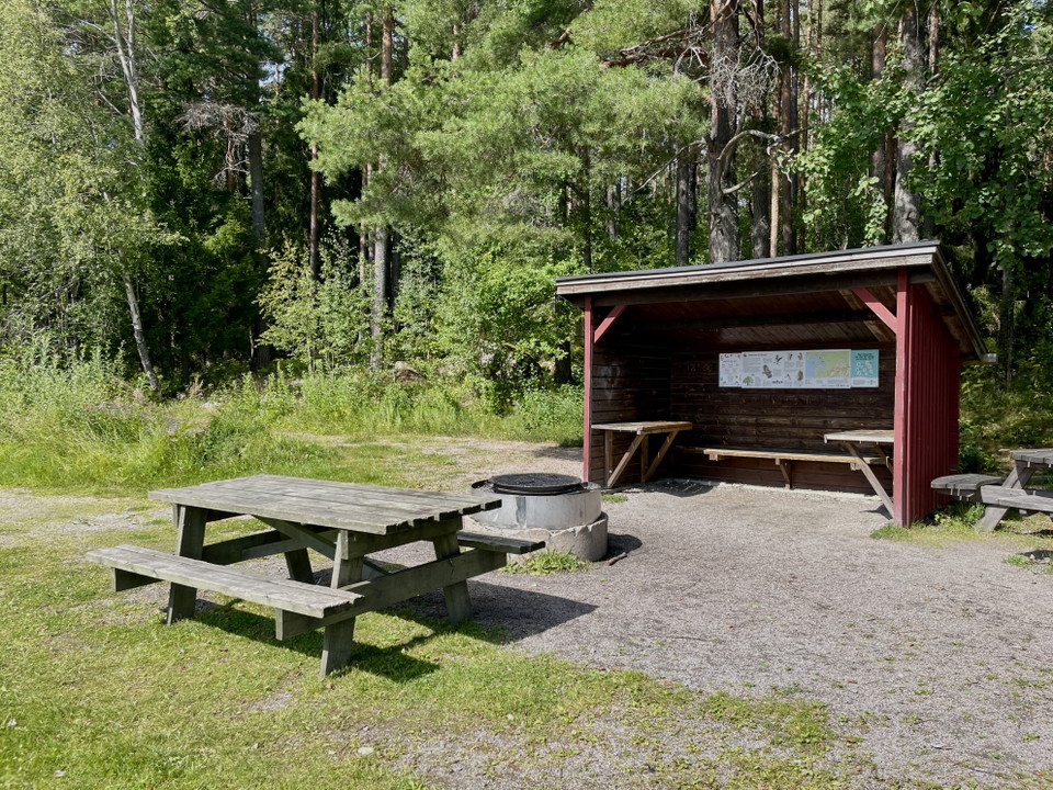 Vindskydd med bänkar längs sidorna, bänkbord och grillplats framför.