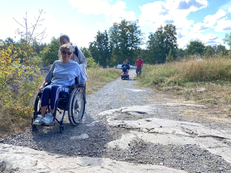 En kvinna i rullstol med assistent, bergigt underlag.