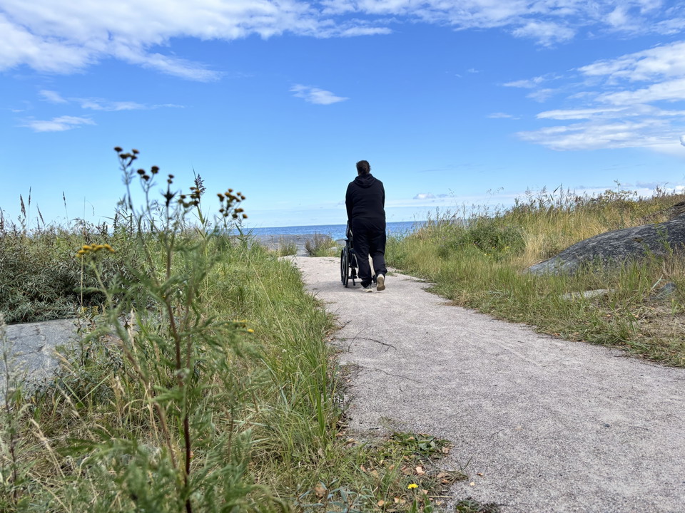 Två kvinnor varav en i rullstol promenerar längs en grusad gångväg.