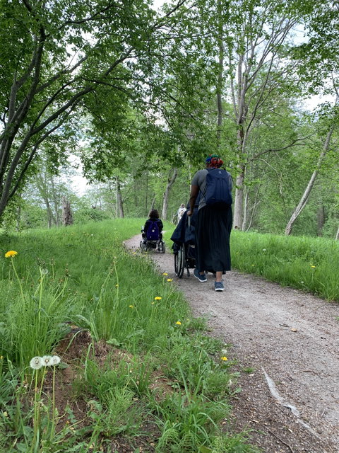 Två personer med rullstol och en assistent promenerar längs smal grusväg i naturen.