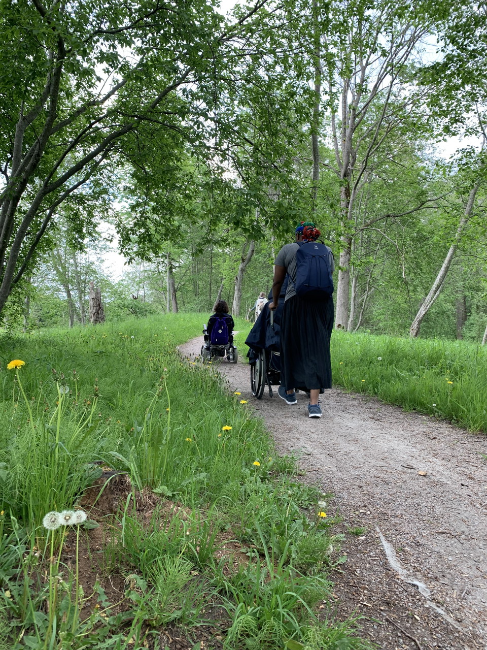 Två personer med rullstol och en assistent promenerar längs smal grusväg i naturen.