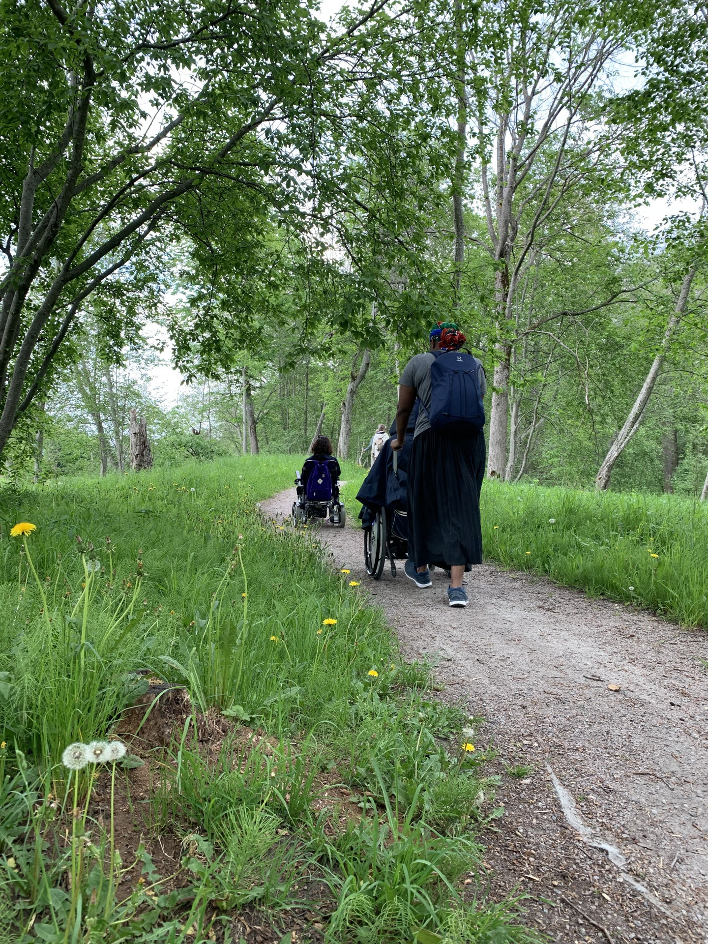 Två personer med rullstol och en assistent promenerar längs smal grusväg i naturen.