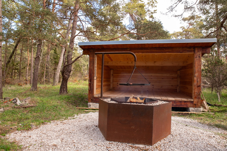 Ett vindskydd i trä med grillplats framför i skogsmiljö.