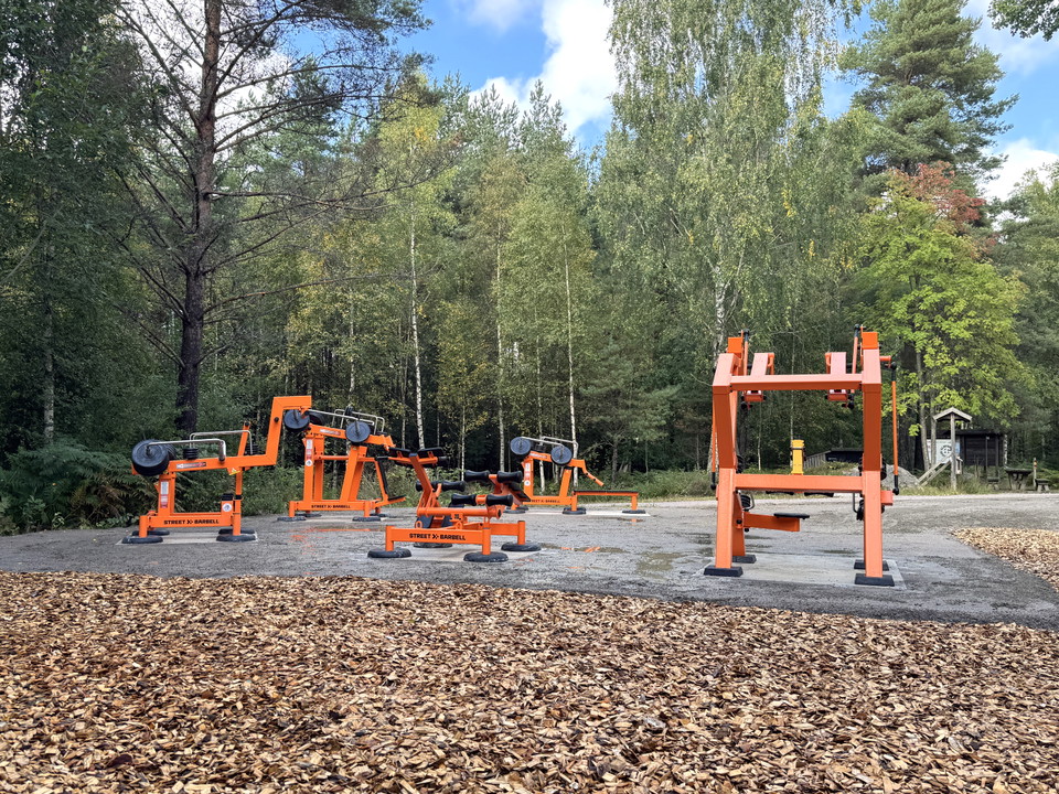 Five orange metal exercise machines are located outdoors. Trees can be seen in the background.