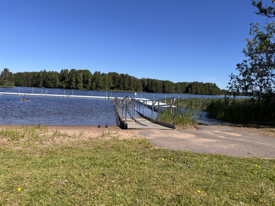 Gräsmatta, lite sandstrand, en ramp som leder ner i vattnet och bryggor.