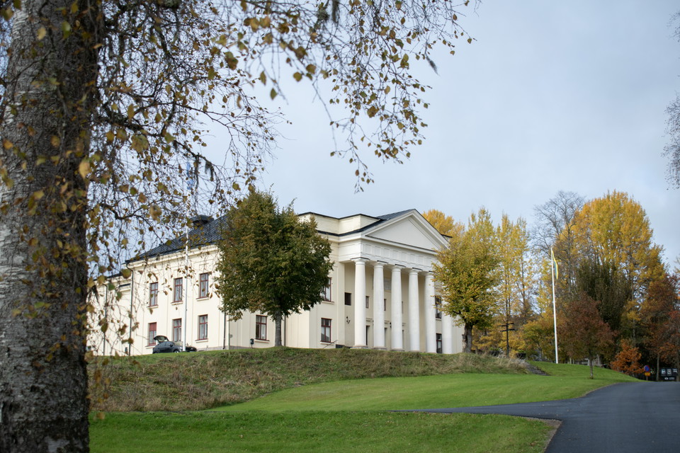 Ståtlig byggnad i engelskt sekelskiftesstil med människor sittandes på bänkar vid entrén. 