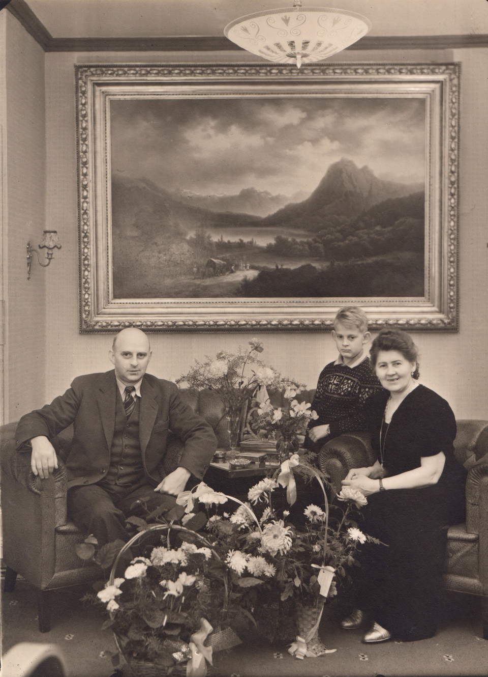 Leonard och Sigrid sitter i varsin fåtölj, emellan dem står massa blombuketter och bakom dem hänger en stor tavla. Jarl står snett bakom sin mor.