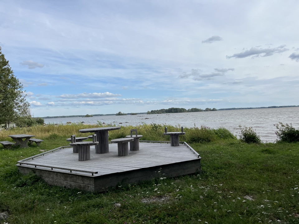 En upphöjd träterrass med bort och sittplatser i gräset vid vatten.