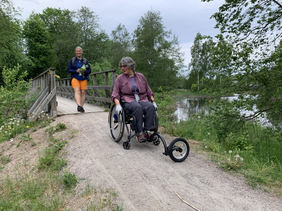 Kvinna i rullstol på grusväg precis efter liten bro.