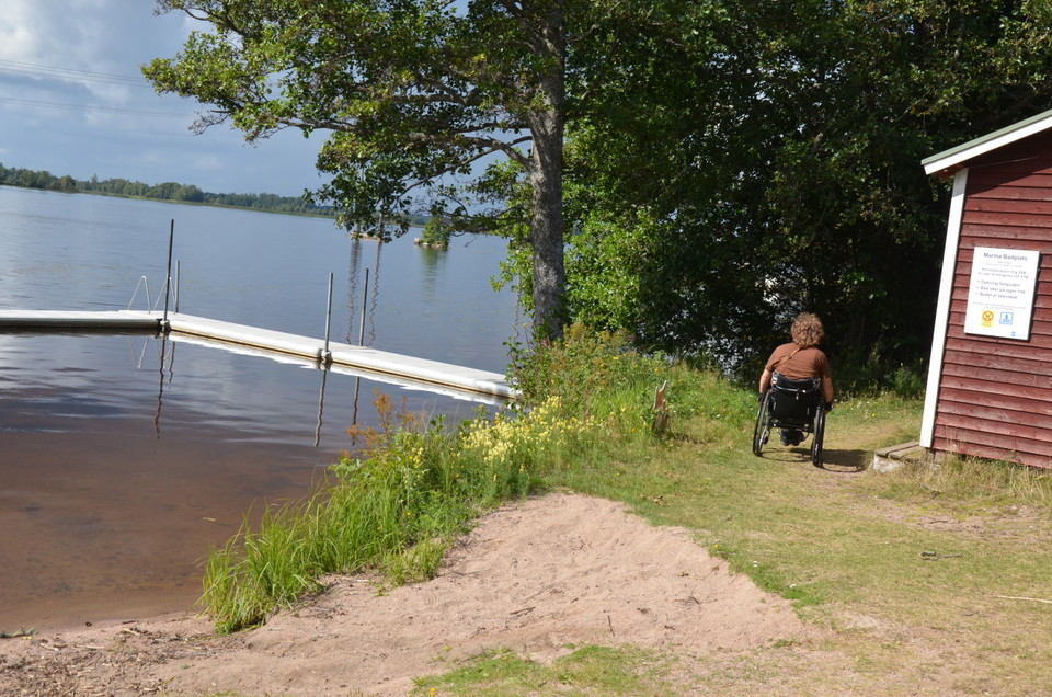 Sandstrand med brygga och en person i rullstol.
