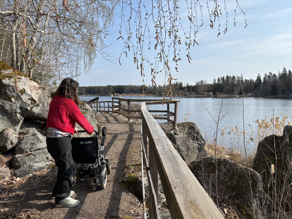 En person skjuter en rullstol längs en grusad gångväg vid en sjö. Längs gångvägen finns ett räcke.