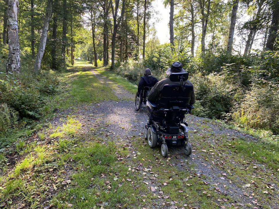En bild på grusväg i skogen med två personer i rullstol.
