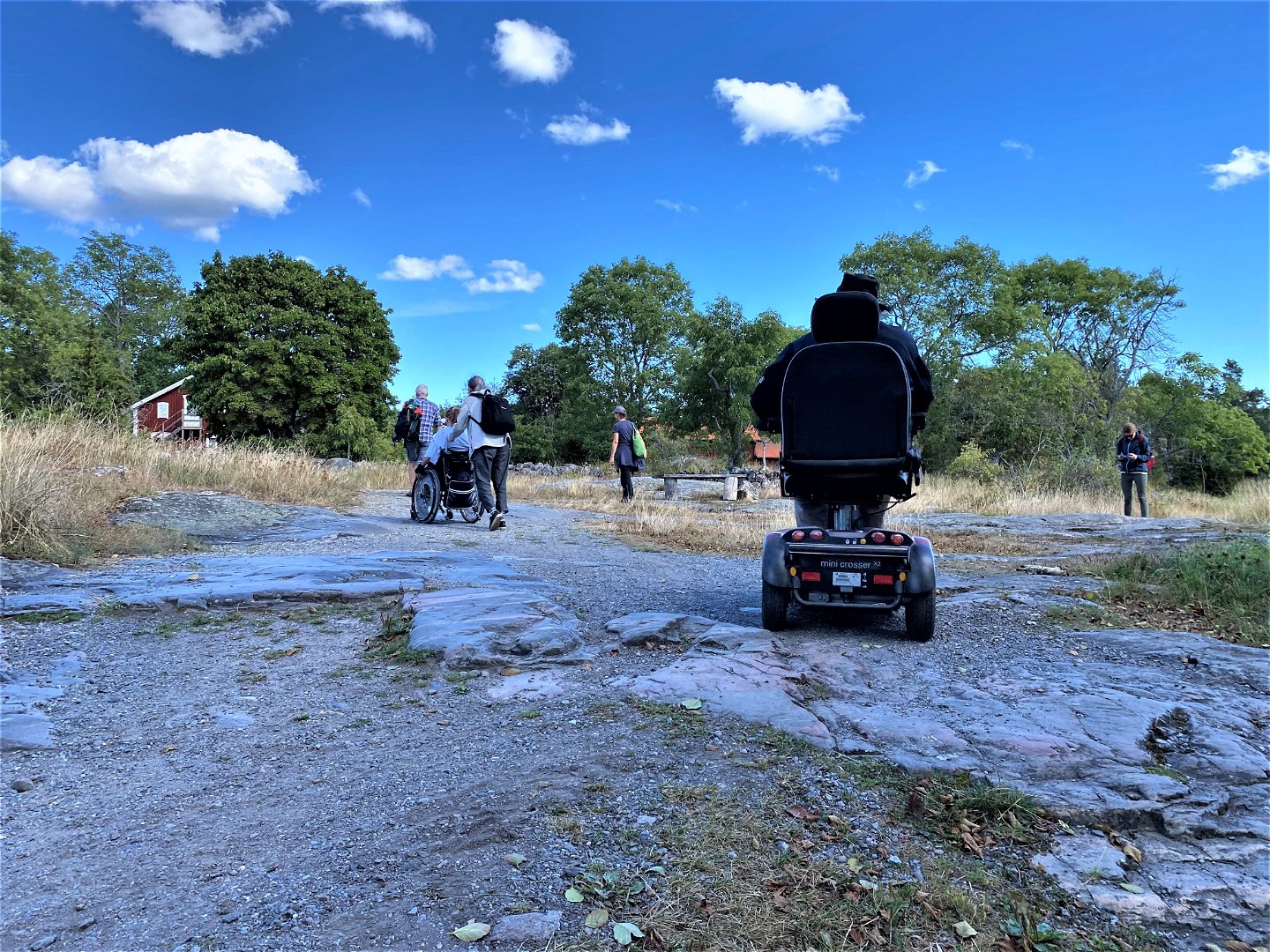 En grupp personer rör sig bortåt på stenig mark. En man kör permobil och en person blir skjutsad i rullstol. 