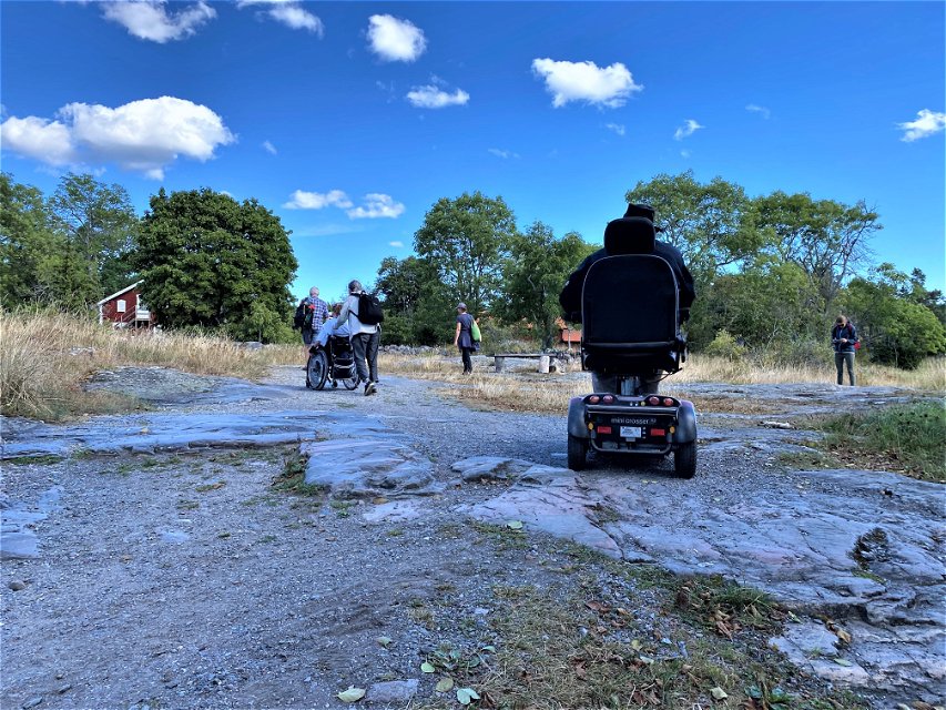 En grupp personer rör sig bortåt på stenig mark. En man kör permobil och en person blir skjutsad i rullstol. 