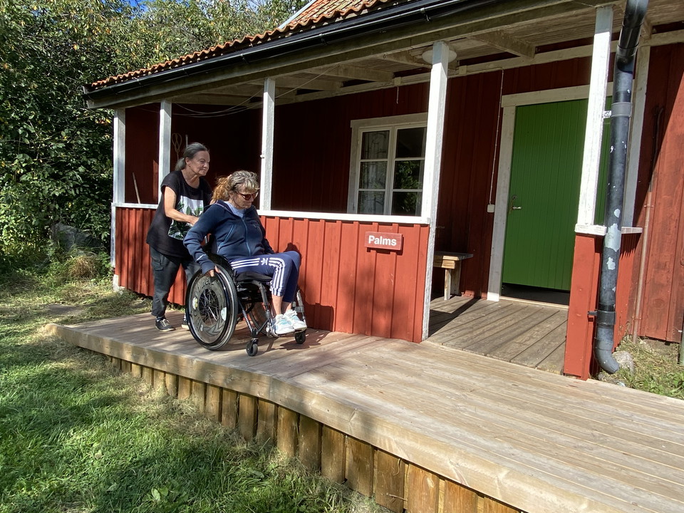 En kvinna i rullstol blir skjutsad uppför en ramp som leder fram till entrén av ett hus. 
