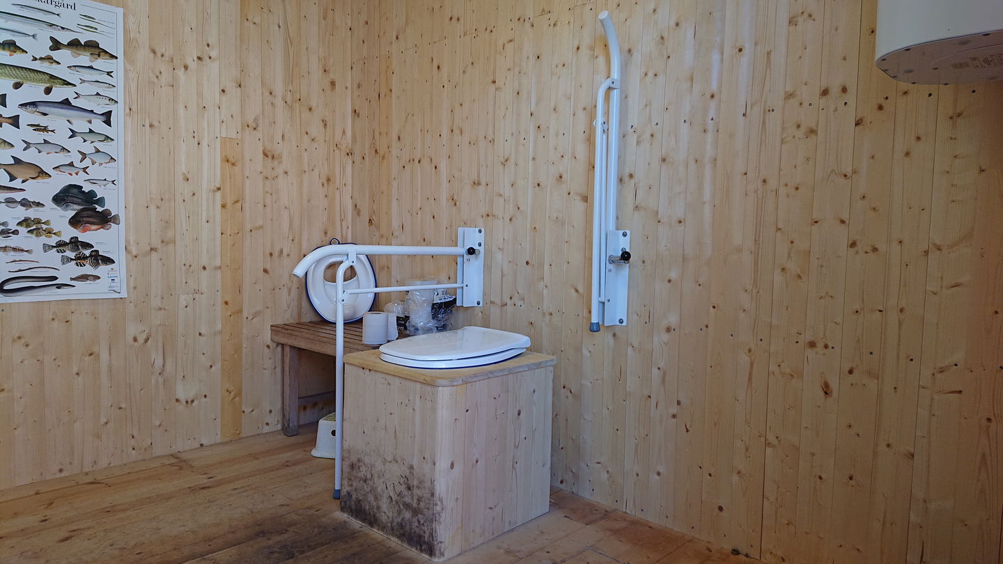 En rymlig torrtoalett med handtag vid toalettstolen. Bredvid står en hylla med toapapper och en fiskplansch hänger på väggen.