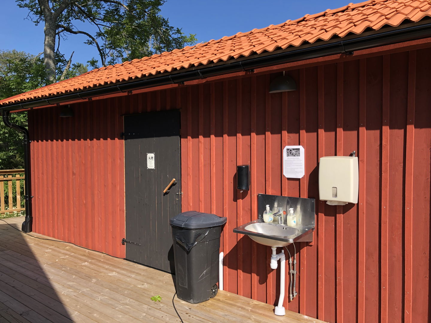 En toalettbyggnad med handfat och en soptunna utanför. Framför ingången är det ett trädäck.