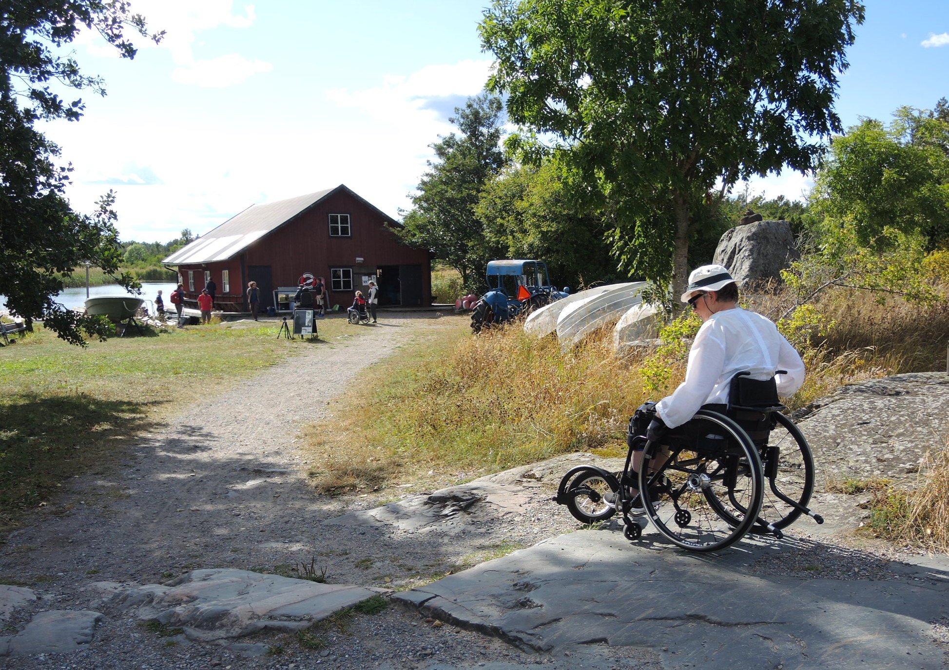 En man tar sig fram i rullstol över stenig mark. I bakgrunden står en byggnad med några människor framför.