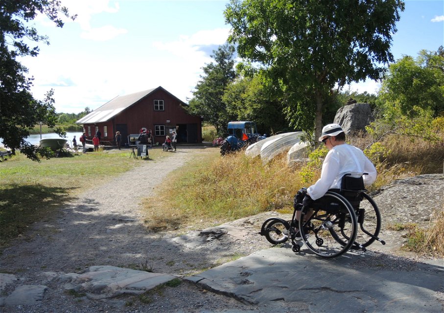 En man tar sig fram i rullstol över stenig mark. I bakgrunden står en byggnad med några människor framför.