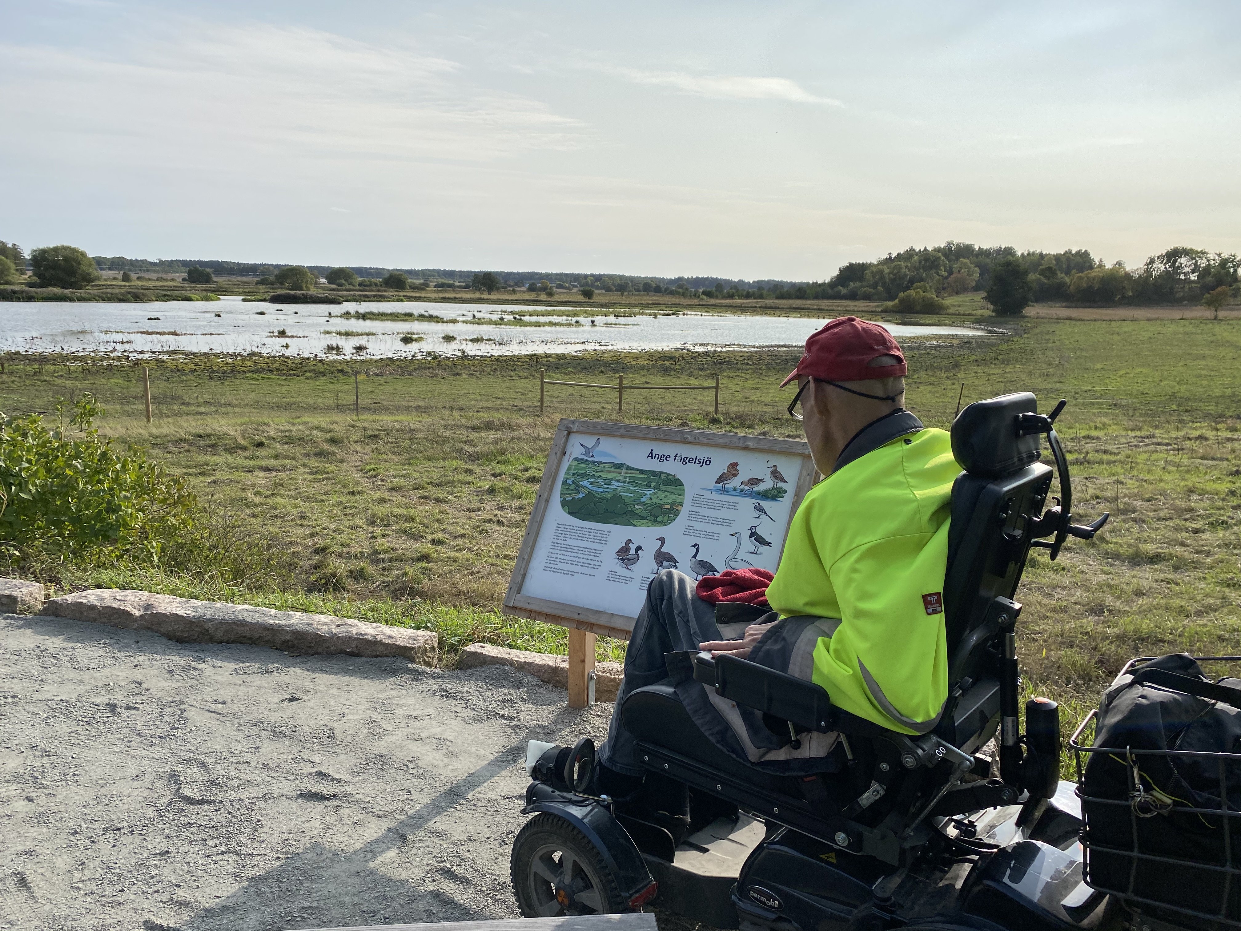 En man i rullstol står på vid en informationstavla på en grusplan. Runt grusplanen finns höga kanter av sten. Framför planen syns våtmark och vatten. 