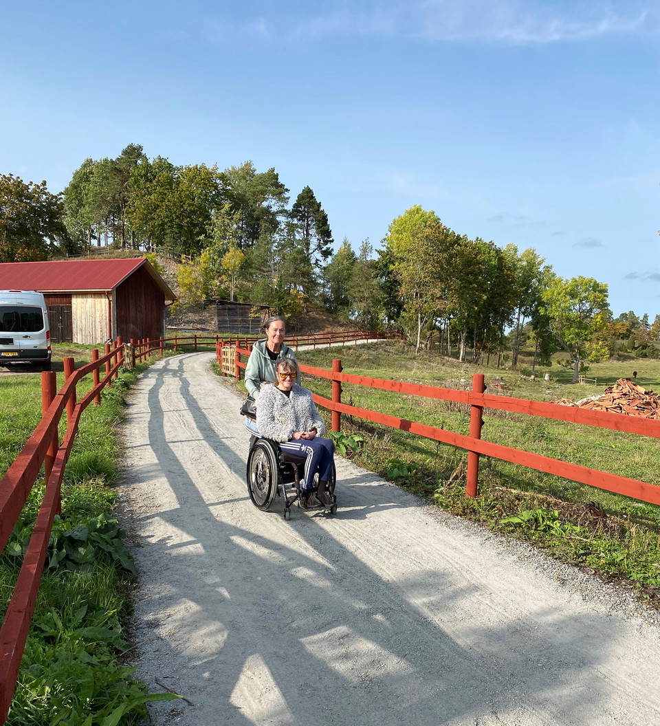 En kvinna skjutsar en man som sitter i rullstol längs en bred grusväg mellan två staket.