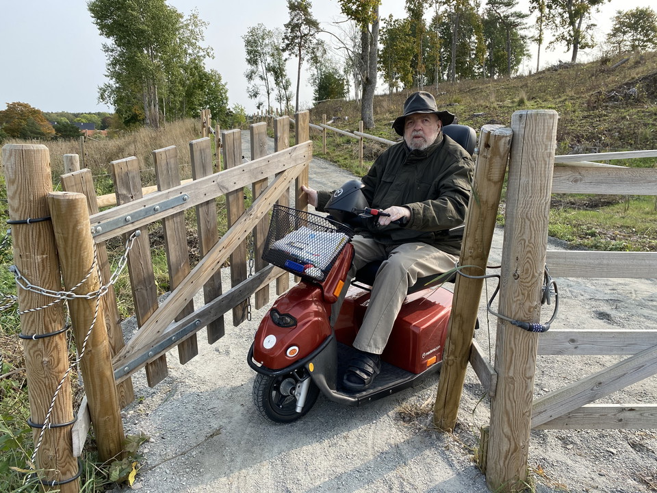En man i permobil öppnar en trägrind  för att kunna passera igenom. Grinden är bred och marken är jämn och grusad.
