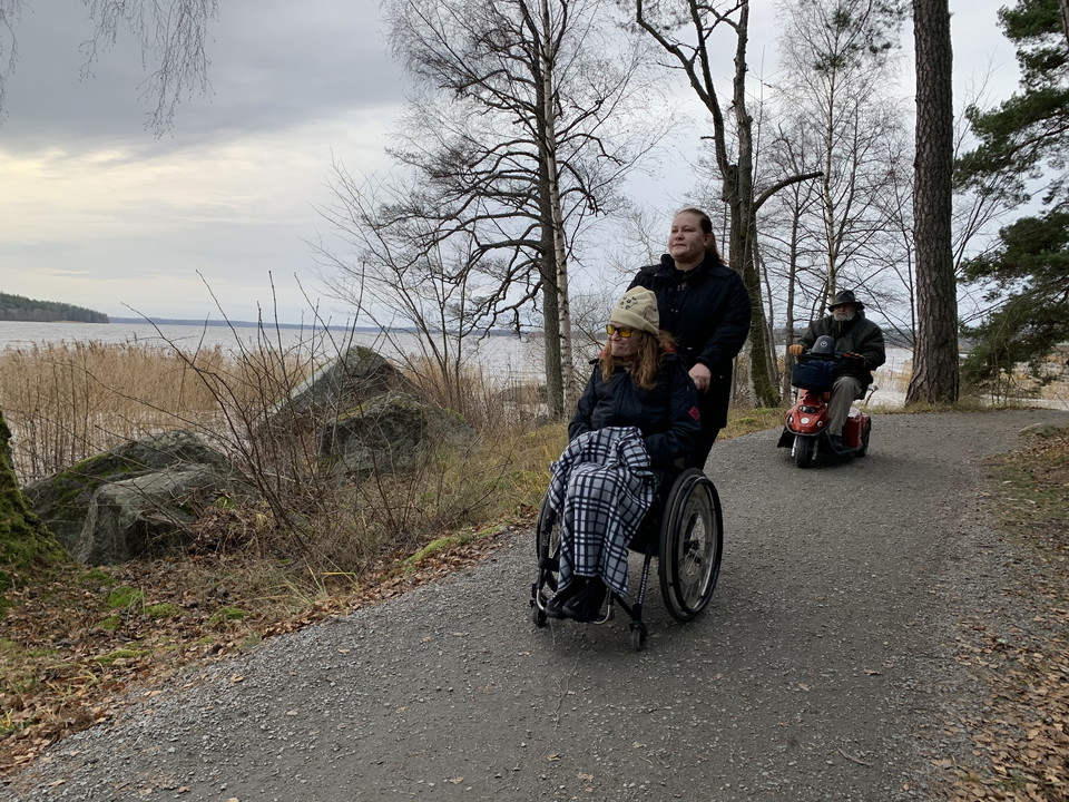 Hårdgjord stig med två personer i rullstol och en ledsagare. Två stora stenar på vänster sida med vatten i bakgrunden.
