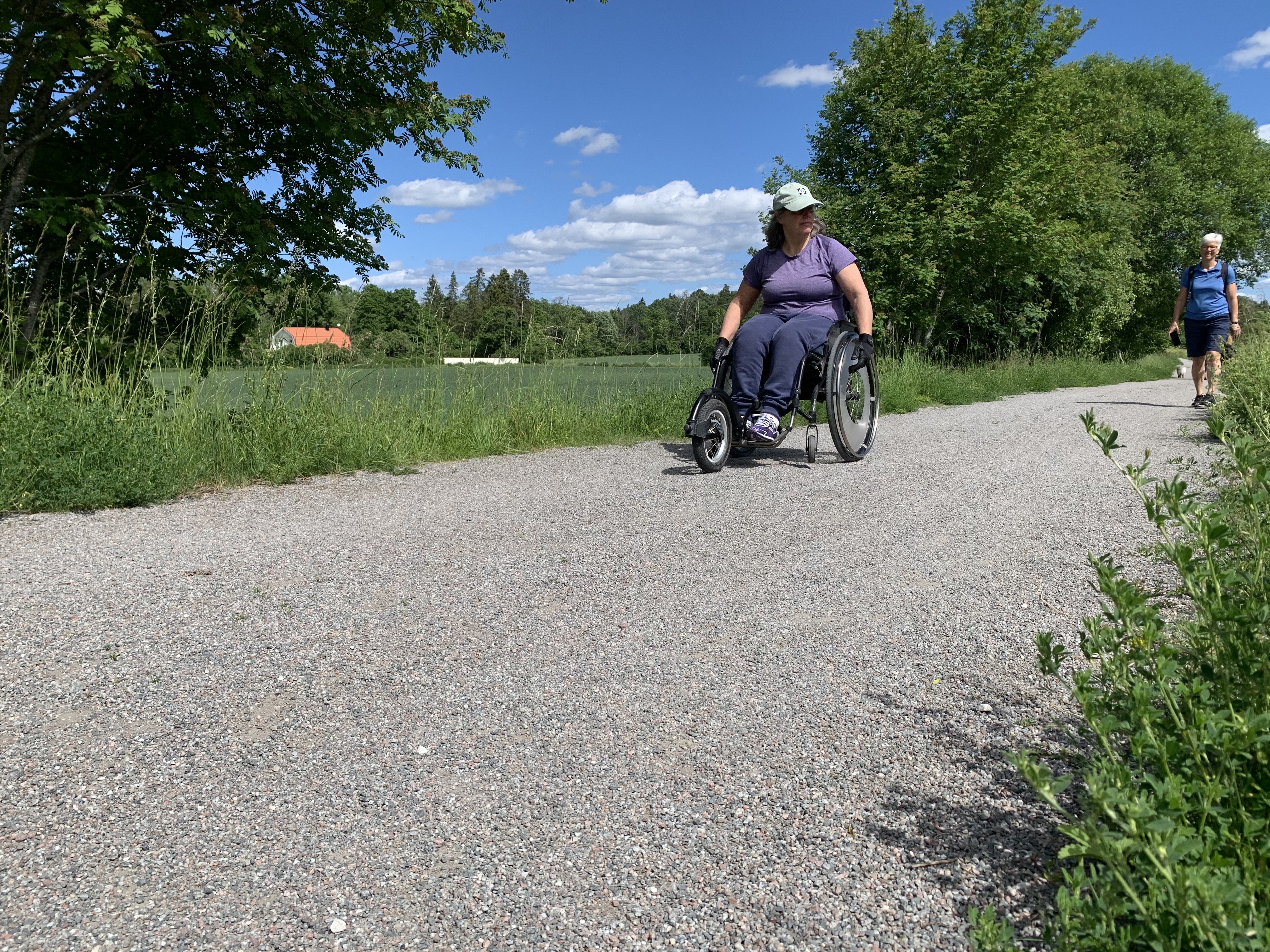 En kvinna i manuell rullstol åker på en bred grusstig som ser jämn ut.