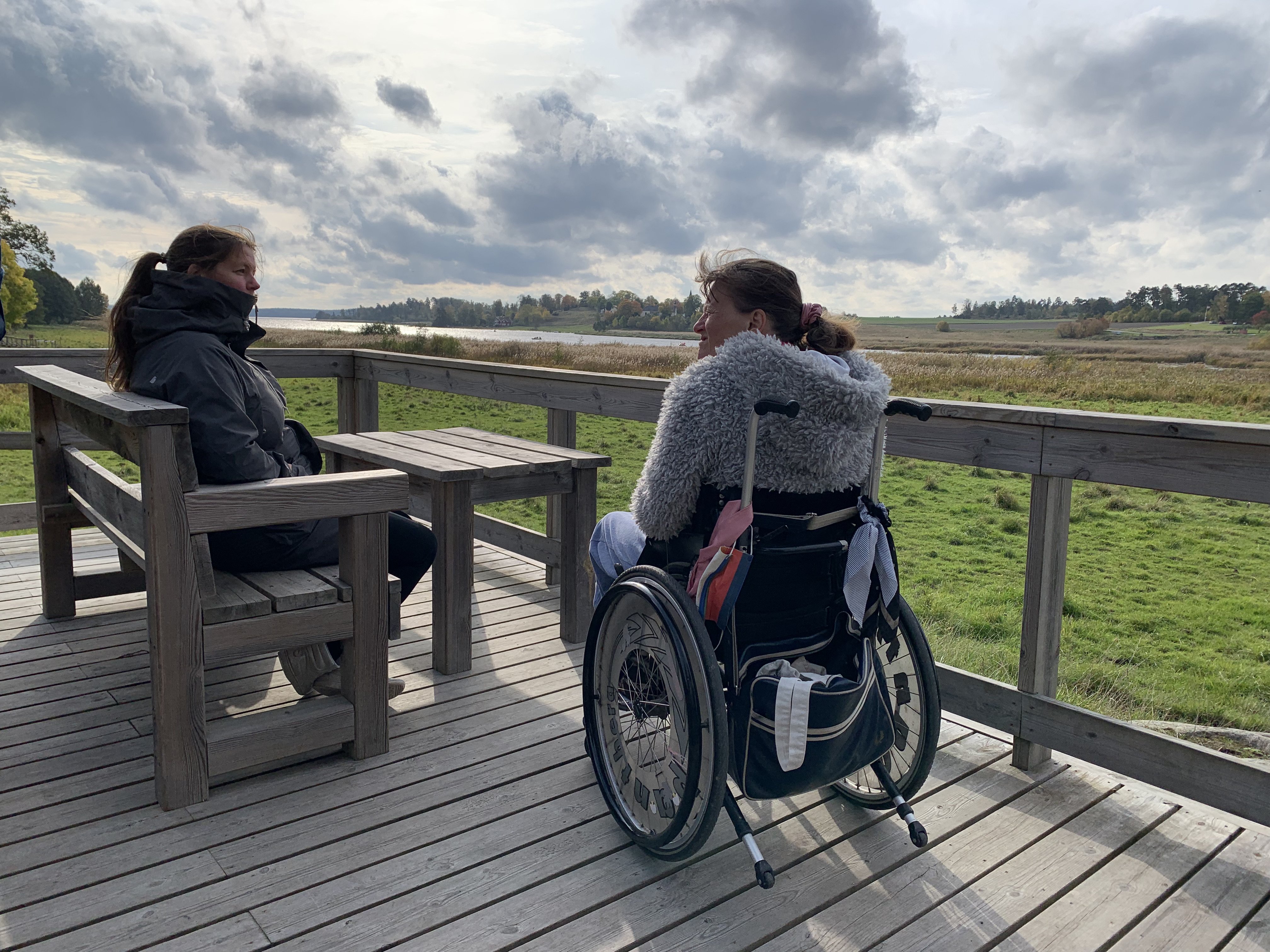 Två kvinnor vid fikabord, den ena sitter i rullstol. Utsikt över öppen mark och en vik