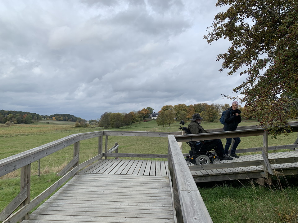 En träspång i naturen som svänger 90 grader. Två personer vandrar, den ena av dem sitter i rullstol.