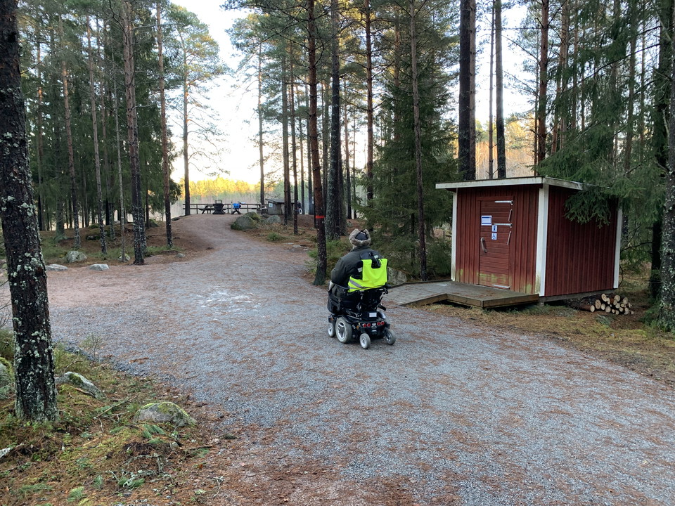 En person som kör rullstol. Dass och rastplats i bakgrunden.