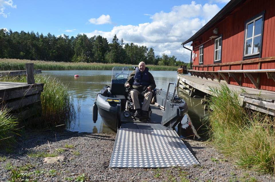 En man i rullstol på en båt med rampen nedfälld. Bryggor och en sjöbod på sidan av båten.