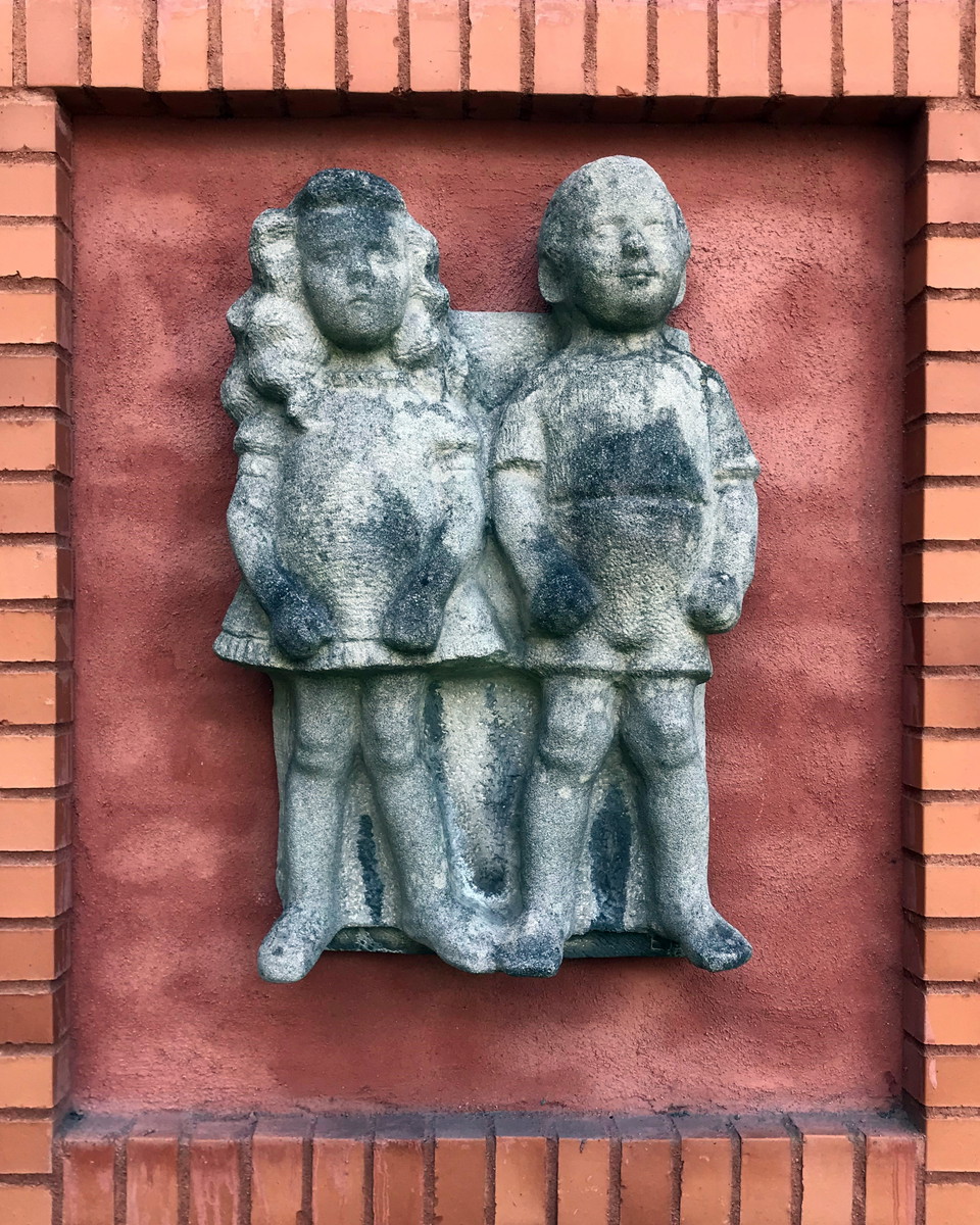 Två grå skulpturer, i form av en flicka och pojke, hänger på rödfärgad vägg.